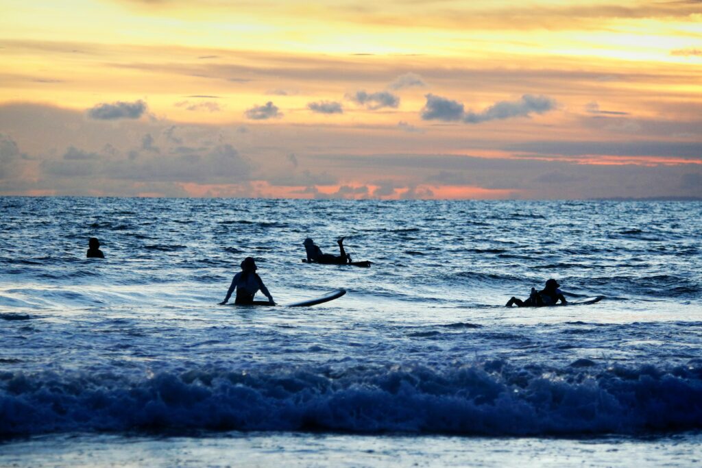 Surfers enjoying sunset waves at Kuta Beach, Bali, with a vibrant sky.